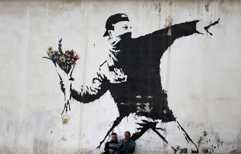 768x492_two-men-are-sitting-in-front-of-a-famous-graffiti-of-british-street-artist-banksy-painted-on-a-wall.jpg
