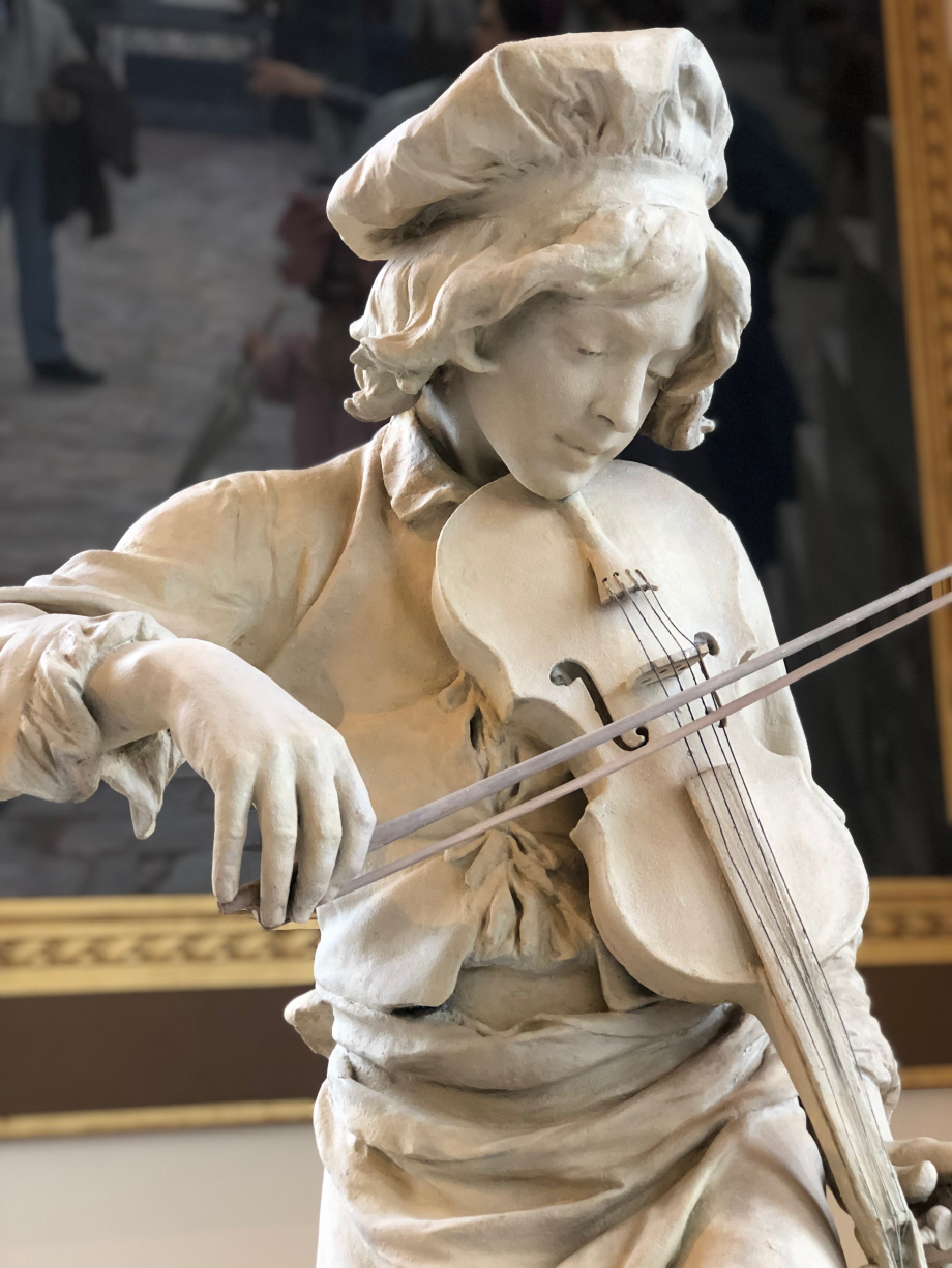 Le sculpteur représente ici Jean-Baptiste Lully (1632-1687), musicien favori de Louis XIV.
Fils d'un meunier de Florence, le jeune Lully est engagé comme garçon de cuisine à la cour de France.
il est ici coiffé d'une toque et vêtu d'un tablier, le pied posé sur une casserole.
L'enfant prodige s'apprête à jouer du violon et à révéler ses dons exceptionnels.